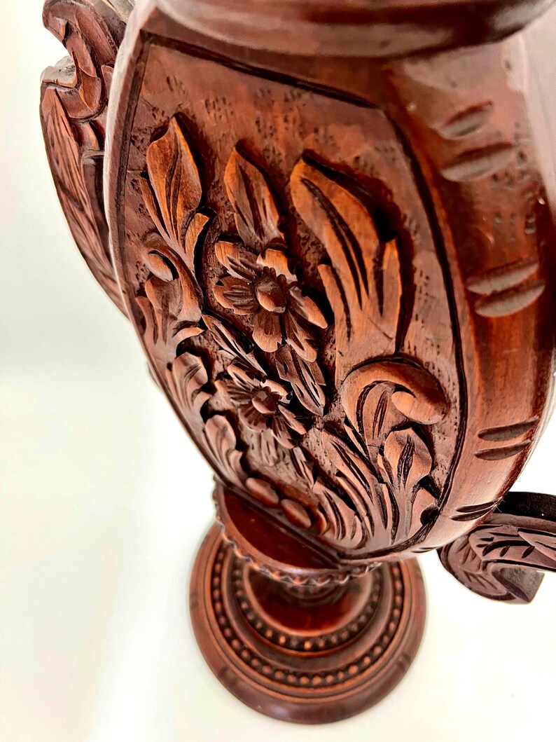 Puede incluir: Primer plano de un objeto decorativo de madera tallada de color marr&oacute;n oscuro. El objeto presenta intrincados dise&ntilde;os florales y de hojas. La base es redonda con un patr&oacute;n detallado. La madera tiene un tono rico y c&aacute;lido, y la talla es muy detallada.