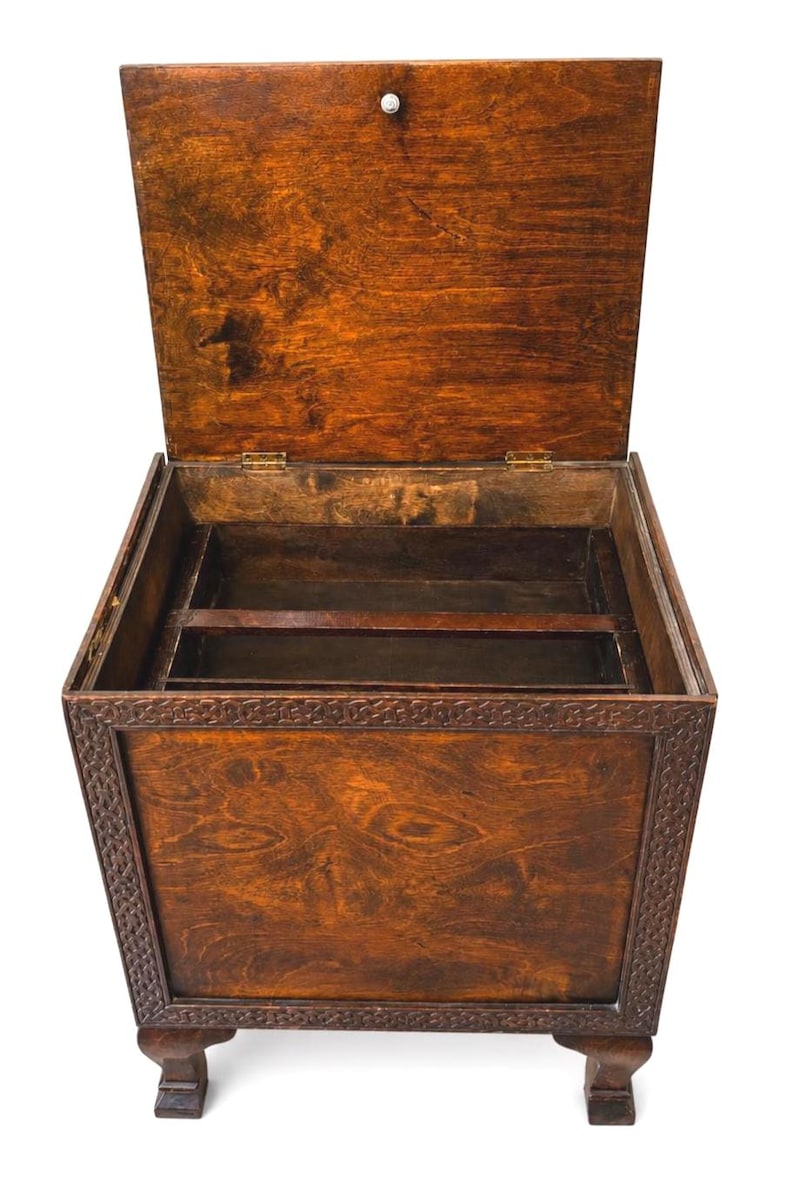 May include: Open wooden chest with a hinged lid, showcasing a rich, warm brown finish. The chest features decorative carved detailing around the edges and rests on short, sturdy legs. The interior reveals compartments, suggesting storage for various items.