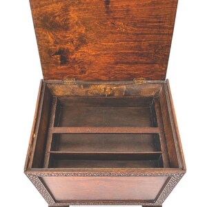 May include: A wooden box with a hinged lid, open to reveal interior compartments. The box is stained a warm brown color, with decorative carvings around the edges. The lid is slightly ajar, showing the interior storage space.