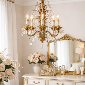 May include: A gold chandelier with crystal accents hangs above a white vanity with gold trim. The vanity holds a mirror, flowers, and perfume bottles. A bouquet of pink roses sits in a vase on the vanity.