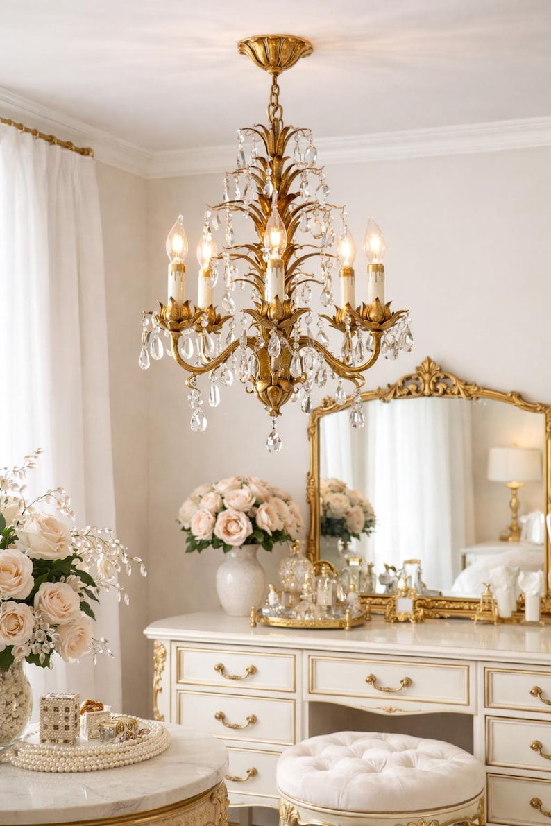May include: A gold chandelier with crystal accents hangs above a white vanity with gold trim. The vanity holds a mirror, flowers, and perfume bottles. A bouquet of pink roses sits in a vase on the vanity.