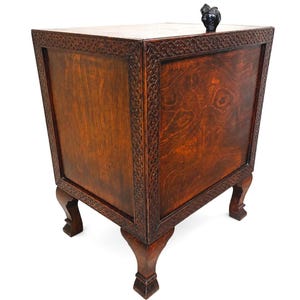 May include: A wooden cabinet with a dark brown finish and intricate carved detailing around the edges. The cabinet has four legs and a small, dark-colored sculpture on top. The wood grain is visible, adding to its rustic charm.