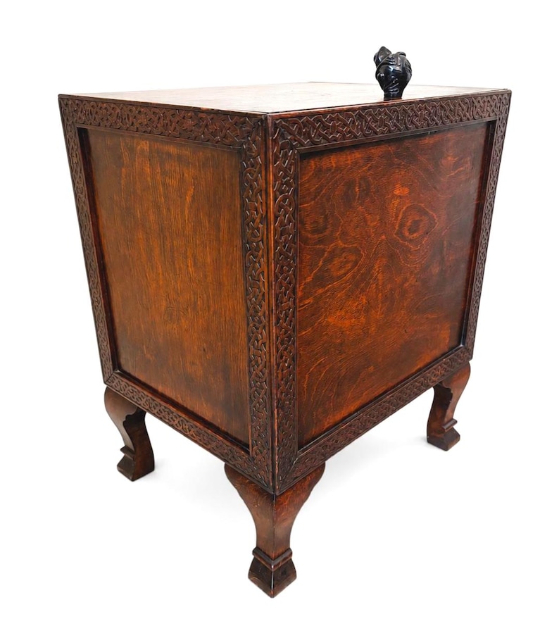 May include: A wooden cabinet with a dark brown finish and intricate carved detailing around the edges. The cabinet has four legs and a small, dark-colored sculpture on top. The wood grain is visible, adding to its rustic charm.