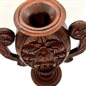 Puede incluir: Un jarr&oacute;n de madera tallada de color marr&oacute;n oscuro con dos asas ornamentadas. El jarr&oacute;n presenta intrincados dise&ntilde;os florales y de hojas, y una amplia abertura. La base es redonda y tambi&eacute;n est&aacute; tallada con un dise&ntilde;o circular. El jarr&oacute;n es probablemente para fines decorativos.