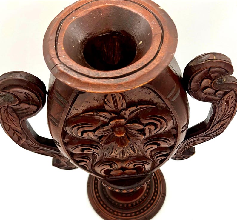 Puede incluir: Un jarr&oacute;n de madera tallada de color marr&oacute;n oscuro con dos asas ornamentadas. El jarr&oacute;n presenta intrincados dise&ntilde;os florales y de hojas, y una amplia abertura. La base es redonda y tambi&eacute;n est&aacute; tallada con un dise&ntilde;o circular. El jarr&oacute;n es probablemente para fines decorativos.