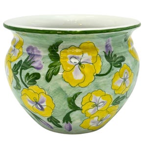 May include: A round ceramic planter featuring a hand-painted design of yellow and light purple pansy flowers with green leaves against a light green brushstroke background. The pot has a wide, flared white rim and a smooth, glossy finish.