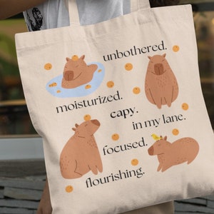 May include: A beige tote bag with a design of capybaras and the text "unbothered. moisturized. capy. in my lane. focused. flourishing."
