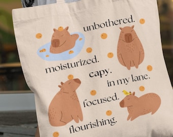 May include: A beige tote bag with a design of capybaras and the text "unbothered. moisturized. capy. in my lane. focused. flourishing."