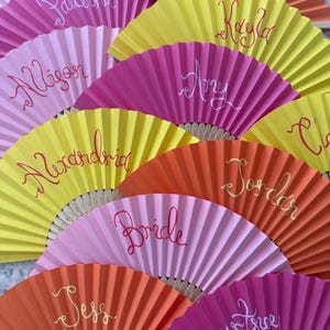 Peut inclure: Une collection d'éventails pliants à main dans des tons vifs de rose, jaune et orange. Chaque éventail porte un nom unique ou le mot Bride écrit en écriture cursive. Les éventails ont des surfaces en papier plissé et des baleines en bois clair.