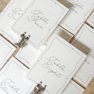 Op de afbeelding: Een verzameling rechthoekige witte papieren tafelkaarten met een dunne zwarte decoratieve rand en elegante cursieve tekst. Elke kaart toont een specifiek tafelnummer, zoals Table Four en Table Eight, vastgehouden door kleine zilveren metalen standaards.