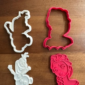 Può includere: Quattro stampini per biscotti in plastica bianca e rosa a forma di Olaf, Elsa, Anna e un pupazzo di neve. Gli stampini sono su una superficie di legno.
