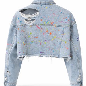 Könnte beinhalten: Hellblaue Jeansjacke mit Used-Details und bunten Farbspritzern. Die verkürzte Jacke hat einen ausgefransten Saum und zerrissene Stellen am Rücken und an den Ärmeln. Ein stilvolles, künstlerisches Oberbekleidungsstück.