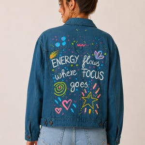 Könnte beinhalten: Eine mittelblaue Jeansjacke mit handgemalten Designs und dem Text "ENERGY flows where FOCUS goes." Auf der Rückseite der Jacke befinden sich ein Sternschnuppen-Motiv, Herzen und andere farbenfrohe, skurrile Elemente.