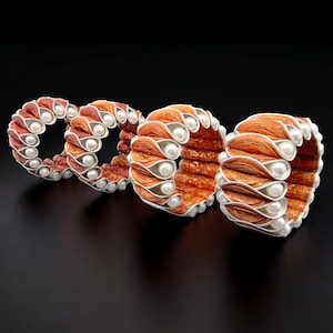 Könnte beinhalten: Drei orangefarbene und silberne Armbänder mit weißen Perlenakzenten. Die Armbänder sind aus gewebtem Material gefertigt und haben ein einzigartiges, strukturiertes Design.