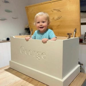 May include: A rectangular wooden toy chest painted in an off-white finish with the name George engraved on the front panel. The chest features a large, hinged lid with a natural light wood grain interior and metal hardware supports on the sides.