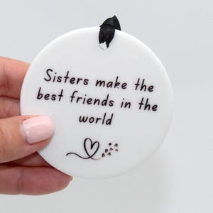 May include: White ceramic ornament with black text that reads "Sisters make the best friends in the world". A small heart and floral design are at the bottom of the ornament.