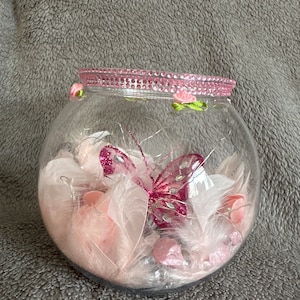 Peut inclure: Un bocal en verre transparent avec un bord en paillettes rose et blanc, rempli de plumes roses, de paillettes et d'un papillon rose pailleté.