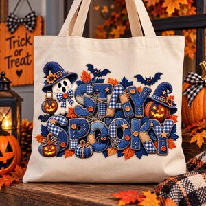 May include: A beige canvas tote bag featuring a decorative applique design with the text STAY SPOOKY. The design includes a white ghost, blue plaid pumpkins, orange jack-o'-lanterns, dark blue bats, and autumn leaves with orange buttons.
