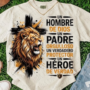 May include: A cream-colored t-shirt featuring a graphic of a lion's head in front of a black cross. The text reads: UN HOMBRE DE DIOS UN PADRE ORGULLOSO UN VERDADERO PROTECTOR UN HÉROE DE VERDAD. The design uses black and gold ink colors.