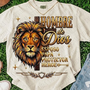 May include: A cream-colored t-shirt featuring a graphic of a lion's head and a cross. The text reads: HOMBRE de Dios, ESPOSO, PAPÁ, PROTECTOR, HÉROE. The design includes gold-toned accents, a key, and a laurel wreath with a shield at the bottom.