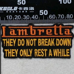 May include: A rectangular black embroidered patch featuring the orange text Lambretta above the yellow text THEY DO NOT BREAK DOWN THEY ONLY REST A WHILE. The patch is positioned next to a black metal ruler showing a width of 3.5 inches.