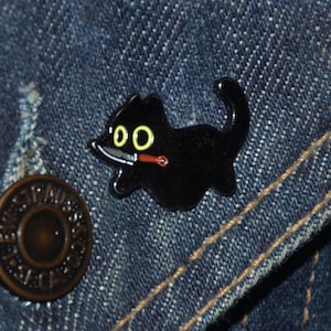 May include: Black enamel pin of a cartoon cat with yellow eyes holding a knife in its mouth. The cat is on a denim background.