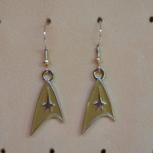 Könnte beinhalten: Ein Paar goldfarbene Ohrringe mit dem Starfleet-Abzeichen aus Star Trek. Die Ohrringe haben eine dreieckige Form mit einem Stern-Emblem in der Mitte und sind an einem silbernen Haken befestigt.