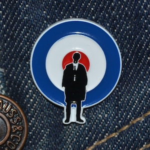 May include: A round enamel pin with a blue and white target design, featuring a black silhouette of a person in a suit. The pin has a red center and a white border. The pin is set against a blue denim background.