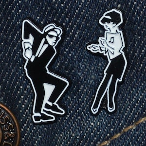 May include: Two black and white enamel pins showing stylized dancing figures. One is a man in a suit and sunglasses, the other a woman holding sheet music. The pins are set against a blue denim background.
