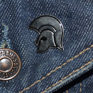 May include: A silver-coloured metal pin shaped like a helmet with a black outline. The pin is on a blue denim background.