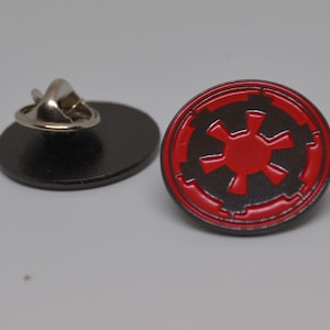 Pode incluir: Um broche de esmalte vermelho e preto com o símbolo imperial de Star Wars. O broche tem um verso de metal prateado e um fecho de segurança.