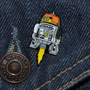 Pode incluir: Um broche de esmalte amarelo, laranja, branco e verde de um robô com um jetpack. O robô tem uma cabeça grande com um único olho e um corpo pequeno com um jetpack.
