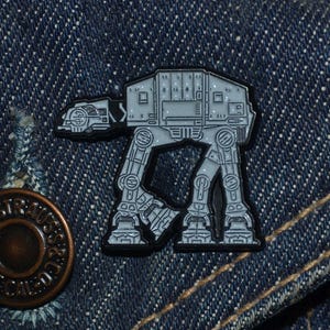 Op de afbeelding: Een grijze en zwarte AT-AT Walker-pin, een voertuig uit de Star Wars-franchise, wordt getoond op een spijkerjasje. De pin is gedetailleerd met de mechanische kenmerken van het voertuig en heeft een zwarte rand. Een messing knoop is zichtbaar linksonder.