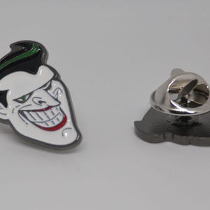 May include: A metal pin featuring a cartoon image of the Joker from Batman. The Joker is depicted with green and black hair, a white face, and a red smile. The pin has a silver metal back with a safety clasp.