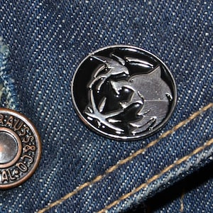 May include: A silver metal pin with a black and white design of a wolf and a bird. The pin is on a blue denim background.