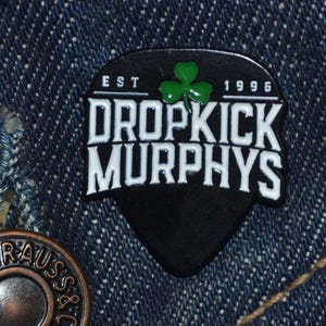 May include: A black and white Dropkick Murphys patch featuring a green shamrock and the text "EST 1996". The patch, shaped like a guitar pick, is set against a denim background.