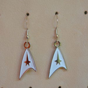 Pode incluir: Par de brincos Star Trek brancos e prateados. Cada brinco apresenta um emblema esmaltado branco com uma estrela dourada e uma borda prateada. Os brincos pendem de ganchos de orelha prateados.