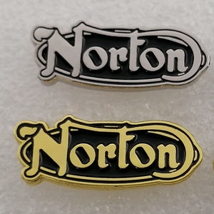 Może przedstawiać: Dwa metalowe szpilki z napisem "Norton" w stylizowanym kroju pisma. Szpilki są czarne z srebrnymi i złotymi literami.