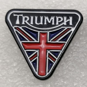 Könnte beinhalten: Ein schwarzer und weißer dreieckiger Pin mit dem Wort "TRIUMPH" in weißen Buchstaben. Die Mitte des Pins zeigt ein rotes Kreuz mit einem blau-weißen Union-Jack-Design.
