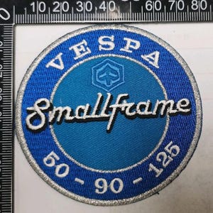 Könnte beinhalten: Ein kreisförmiger gestickter Aufnäher mit einem blau-hellblauen Design und dem weißen Text "VESPA", "Smallframe" sowie "50-90-125". Der Aufnäher liegt neben einem schwarzen Lineal, das einen Durchmesser von etwa 9 cm anzeigt.