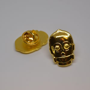 Può includere: Spilla in metallo dorato con una rappresentazione cartoon della testa di C-3PO di Star Wars. La spilla ha una chiusura di sicurezza sul retro.