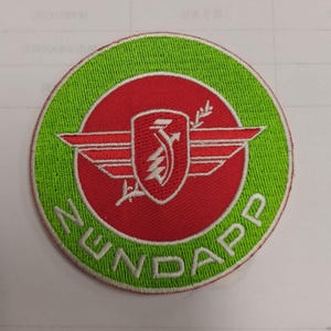 May include: Circular embroidered patch with a green outer ring and a red central design. The design includes a shield, wings, and the word "NUNDAPP" in white. The patch is suitable for clothing.