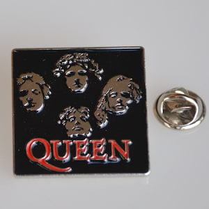 Könnte beinhalten: Ein quadratischer schwarzer Ansteckpin mit dem Wort "QUEEN" in Rot und silbernen Gesichtern der Bandmitglieder. Ein silberner Pin-Rücken befindet sich neben dem Pin. Der Pin ist ein Sammlerstück zum Thema Musik.
