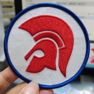 Può includere: Toppa rotonda con un disegno di elmo troiano rosso su sfondo bianco, bordata da un bordo blu cucito. Adatta per abbigliamento o accessori.