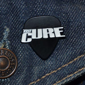 May include: A black guitar pick-shaped pin with the white text "THE CURE" on a denim jacket. The pin is a dark colour and has a glossy finish. A copper-coloured button is visible on the left side of the image.