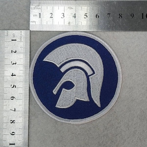 Könnte beinhalten: Gestickter Aufnäher mit einem silbernen Helmdesign auf einem marineblauen kreisförmigen Hintergrund. Der Aufnäher hat einen weißen Rand. Das Design erinnert an einen klassischen Helm. Der Aufnäher hat einen Durchmesser von ca. 7,6 cm.