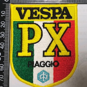 以下が含まれることがあります： 黒いVESPAの文字、大きな黄色のPXの文字、黒いPIAGGIOの文字が刺繍された盾形のワッペン。背景は緑、白、赤の縦縞模様で、下部には小さな青い六角形のロゴがあしらわれています。