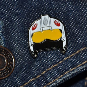 Könnte beinhalten: Ein weißer, schwarzer und gelber Emaille-Pin in Form eines Pilotenhelms. Der Pin hat rote Akzente und ist vor einem blauen Jeans-Hintergrund platziert. Dies ist ein Sammlerstück.