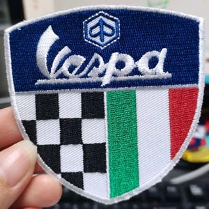 Op de afbeelding: Een schildvormige geborduurde patch met het woord Vespa in witte schrijfletters op een donkerblauwe achtergrond. Daaronder is het ontwerp verdeeld in een zwart-wit geblokt patroon aan de linkerkant en de Italiaanse vlag aan de rechterkant.