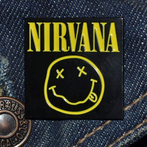 May include: A black square pin with the word "NIRVANA" in yellow block letters above a yellow smiley face with crossed-out eyes and a tongue. Set against a denim background.